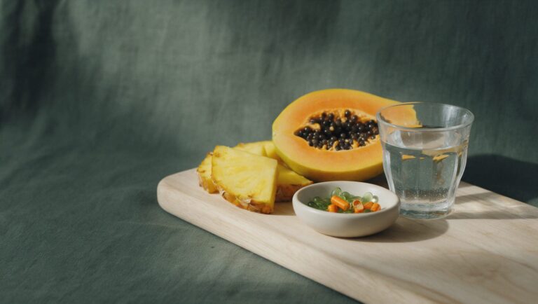 Fresh pineapple and papaya beside a dish of capsules and a glass of water