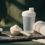 Scoops of creatine and protein powder on a slate board with a shaker bottle and gym towel