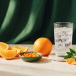 Citrus slices, a dish of orange capsules and a glass of sparkling water on a cream stone surface