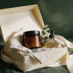 Best Wellness and Recovery Gifts 2026 (by Budget) Open cream gift box with folded linen, an amber jar, a dropper bottle and eucalyptus on green linen